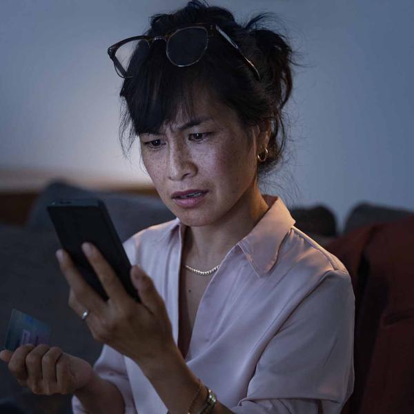 Person sitting on a sofa, holding a smartphone and bank card while looking at the screen in a dimly lit room.